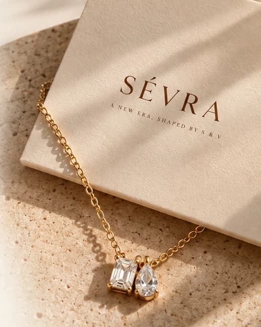 A SÉVRA box and a fine gold chain with a baguette-and-pear pendant resting on travertine in warm daylight